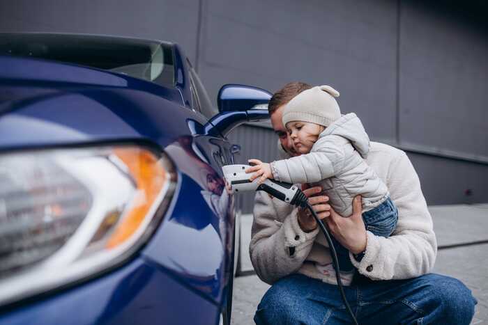 Man holds up young toddler in a knitted hat as she holds the electric charger into the family car